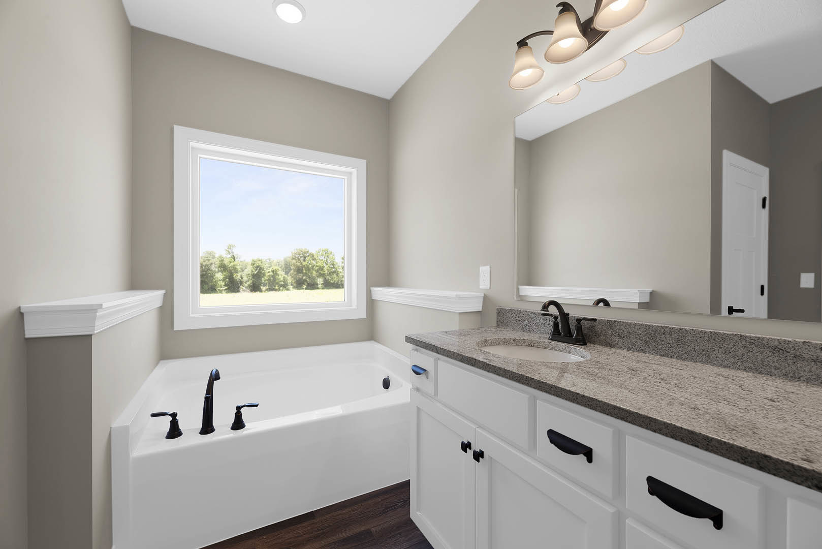 Freestanding bathtub with chrome faucets on wood floor, white sink with black faucet, large window framing leafy trees, white door with black handle, light tile walls and cabinetry