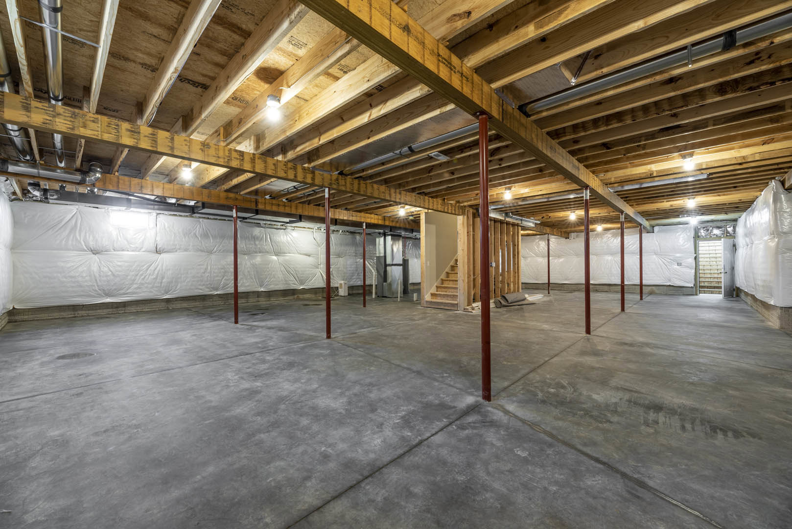 Exposed wooden ceiling beams above a concrete floor with red steel poles, unfinished staircase, large white bag wrapped in plastic against the wall, and white sheet draped in the