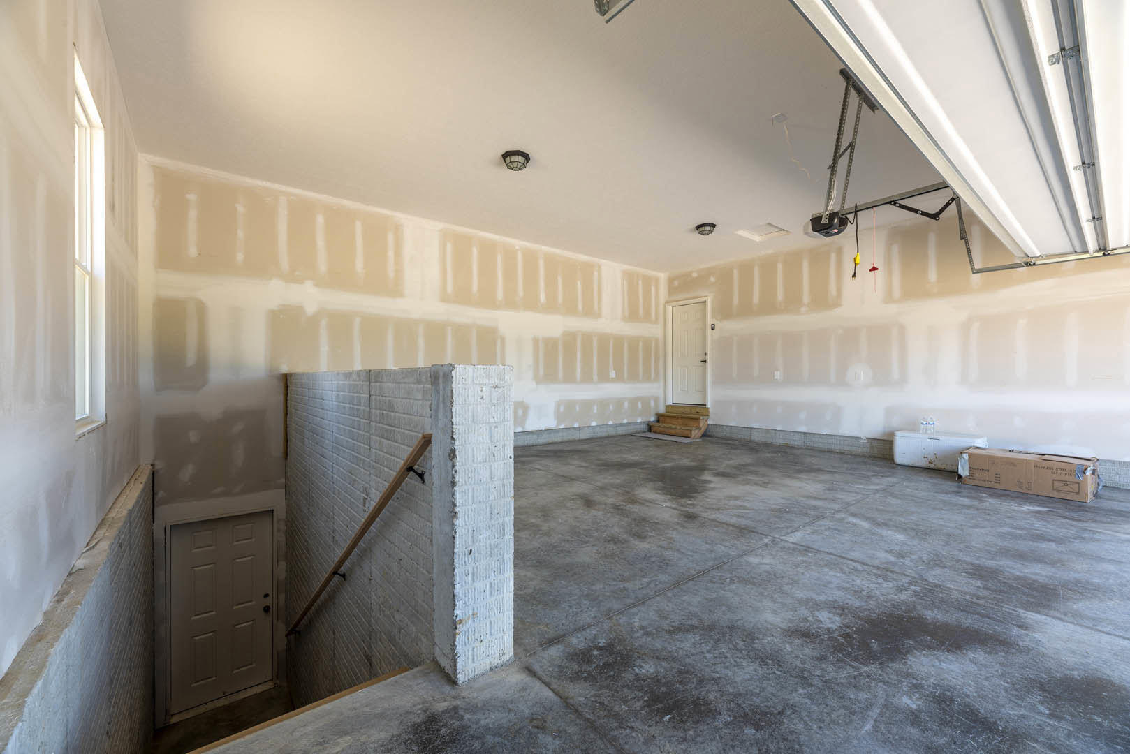 White plaster wall, concrete floor, wooden staircase with matching handrail, white door featuring black handle, brown storage box on grey surface