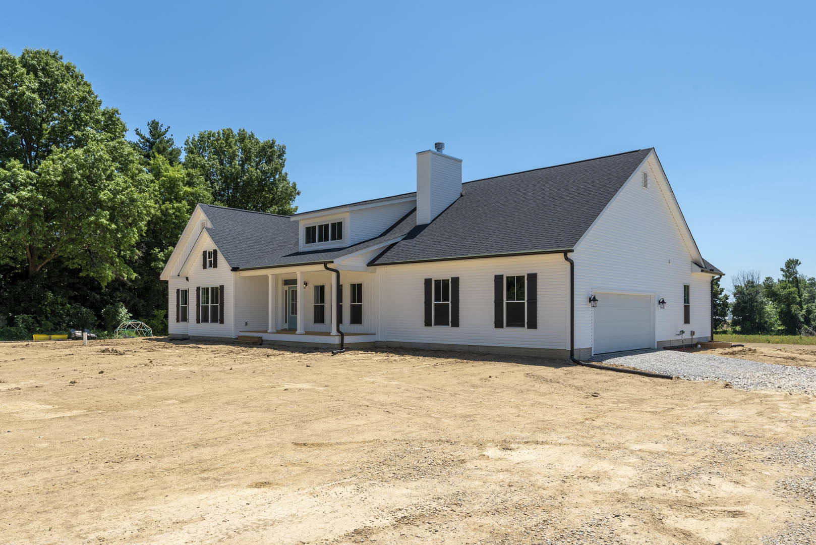 White house with black shutters, white-framed windows, dirt driveway with exposed pipe, leafy tree in front yard