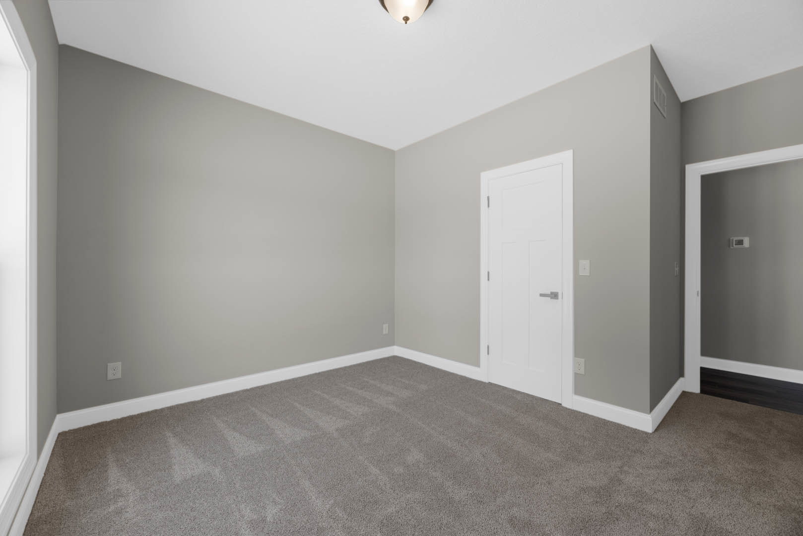 White paneled door with silver handle, grey carpeted floor, white wall with rectangular thermostat, ceiling-mounted light fixture, simple baseboard molding