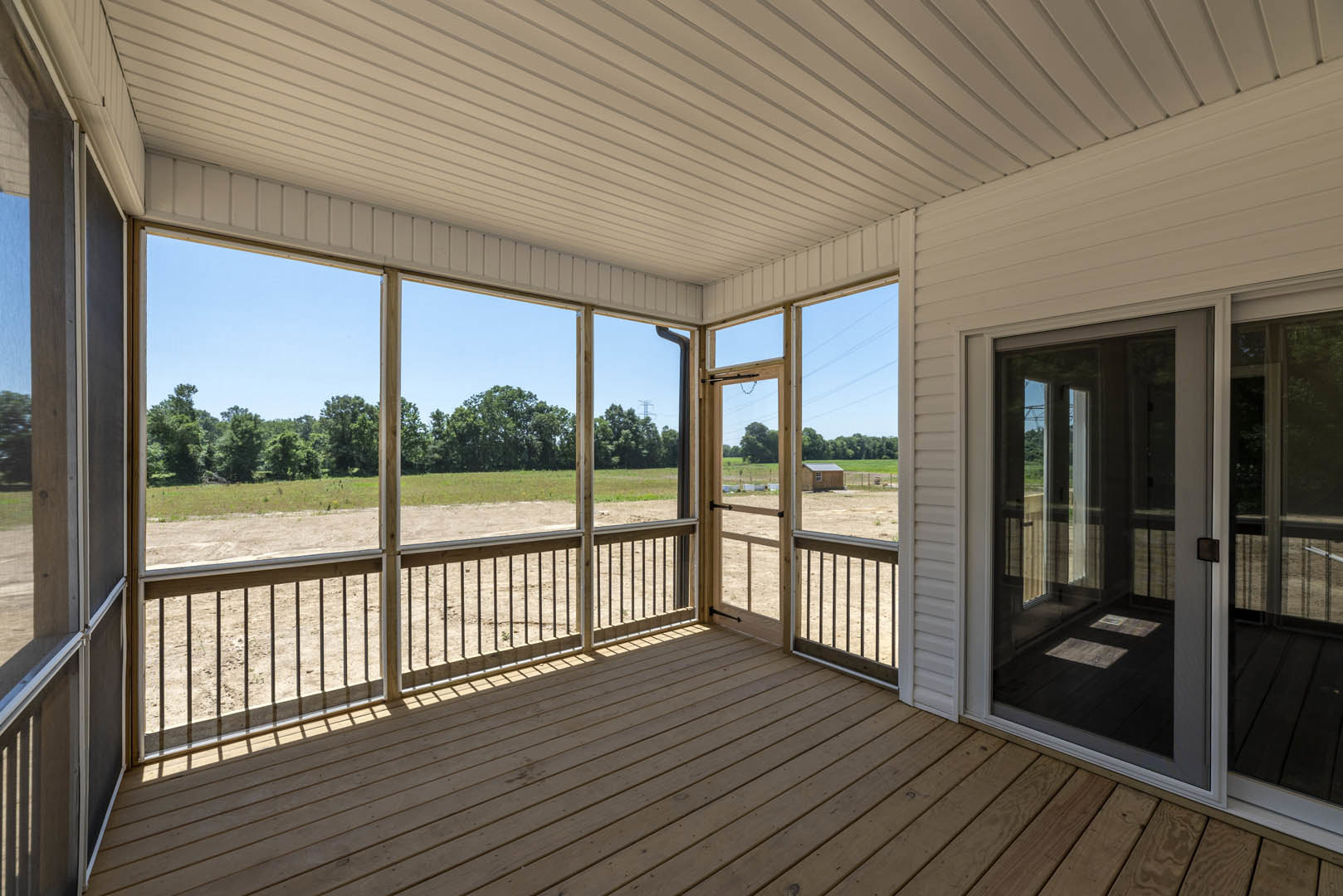Spacious wooden porch with black-accented white ceiling, glass door entry, fenced deck, overlooking open field and distant trees