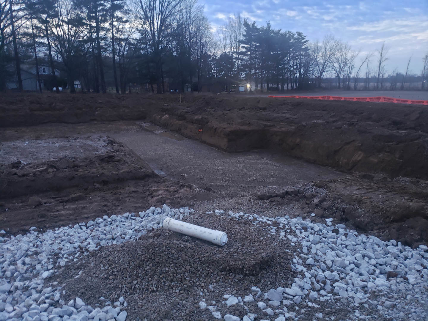 White pipe resting on a pile of gravel at a residential construction site, surrounded by bare trees and cloudy winter sky.