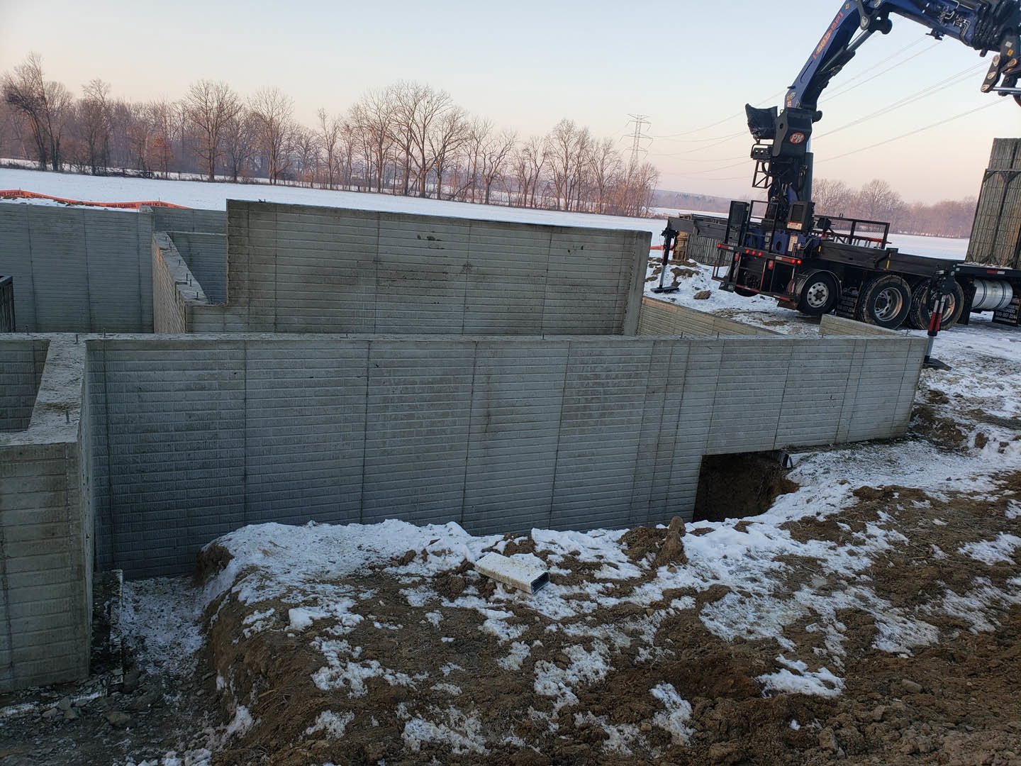 Snow-covered construction site with partially built home, large blue crane truck, white rectangular materials on ground, bare winter trees in background