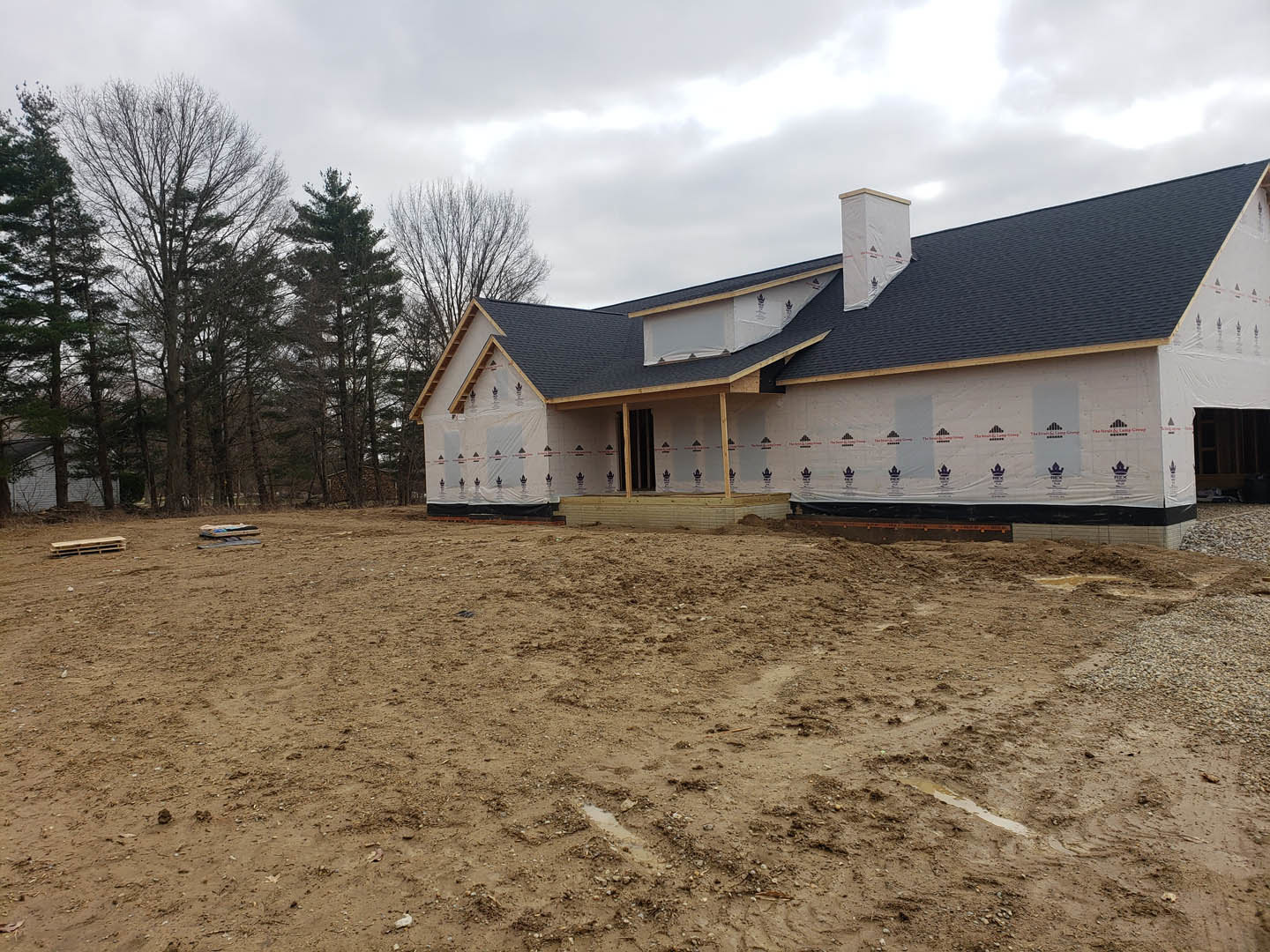 Wood-framed house under construction on a dirt lot, surrounded by leafless trees and cloudy sky