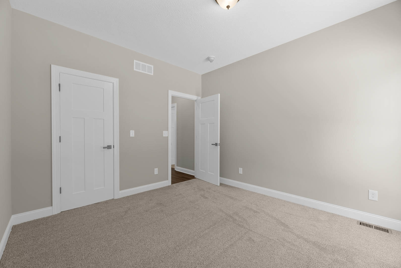 Neutral-toned carpeted room with two white paneled doors featuring silver handles, white walls with base molding, ceiling, and a visible electrical outlet.