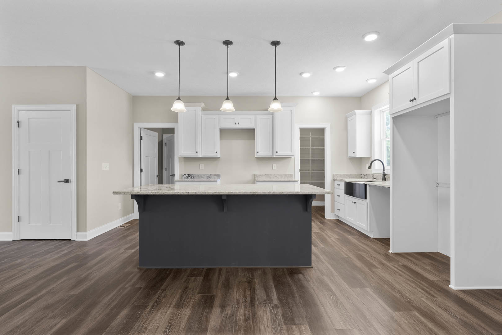 Open kitchen with wood flooring, central island featuring light stone countertop, white cabinetry, stainless steel sink, white door with black handle, recessed ceiling lights, and