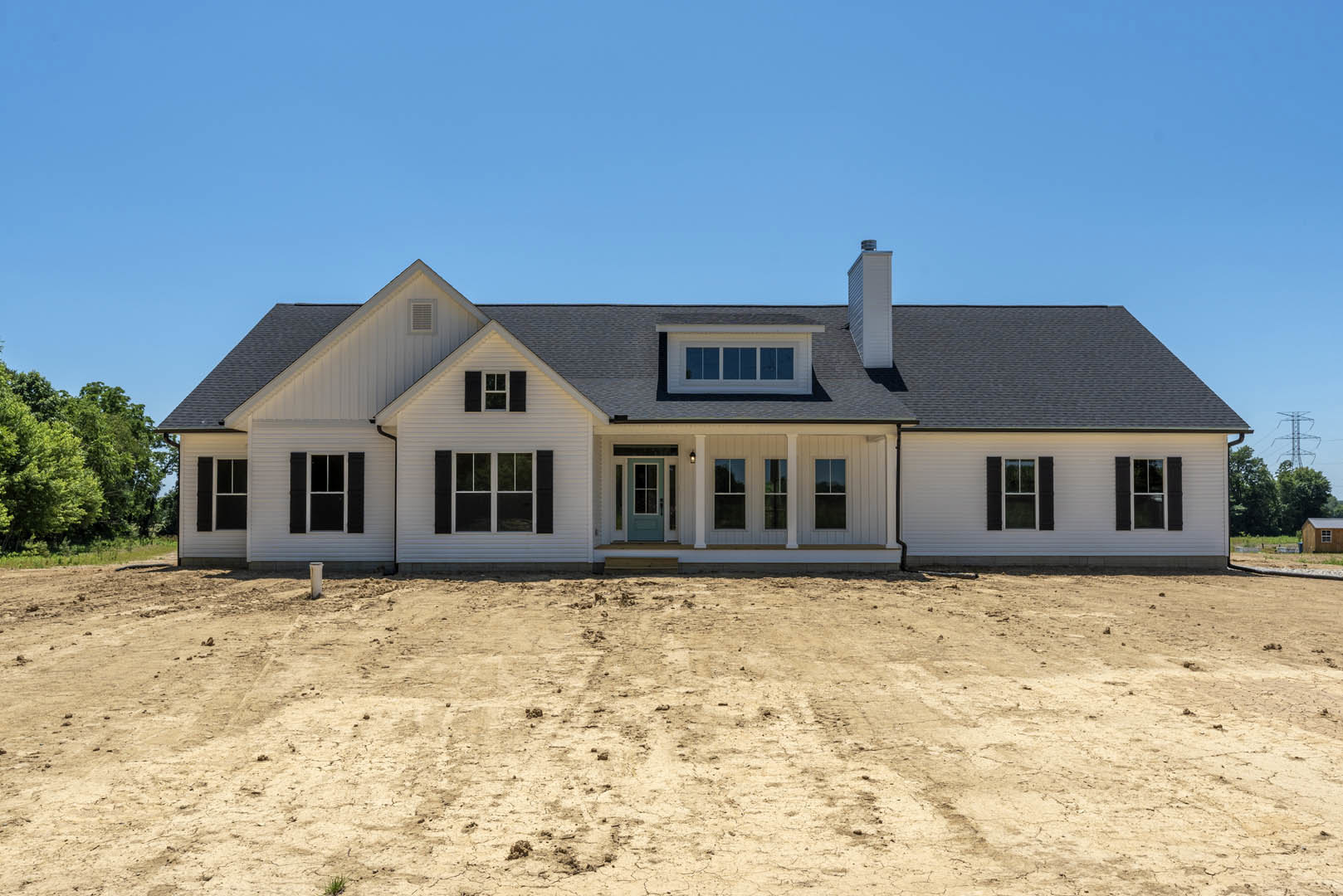 White house with blue door and windows, surrounded by a dirt yard, trees in the background, visible roof and chimney