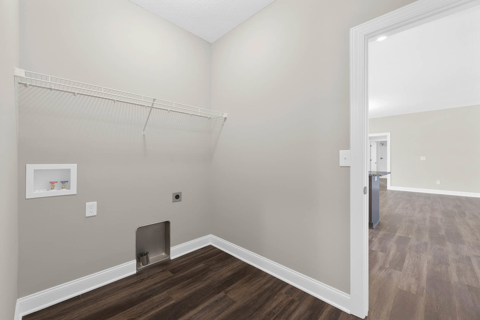 White plaster wall with white electrical outlet and framed plumbing valves, wood flooring with grey surface, white baseboard, metal trash can against wall.