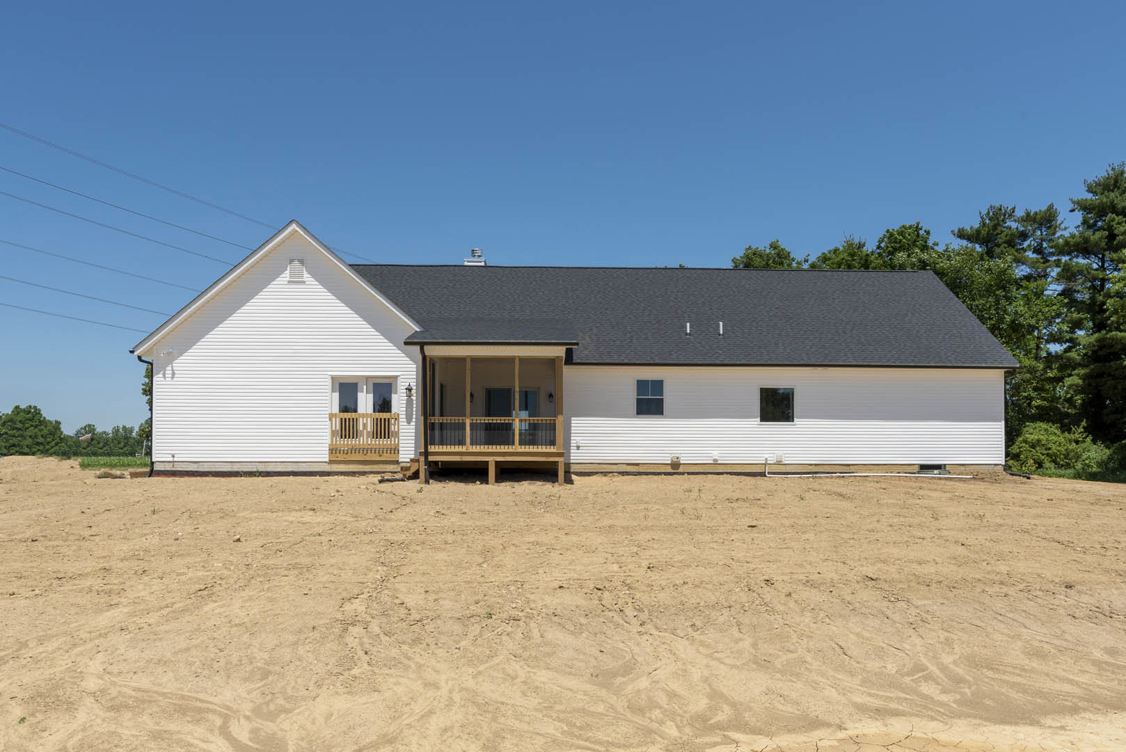 Single-story house with white-framed windows, covered porch featuring black railing and wooden bench, surrounded by dirt field and sparse trees under clear sky