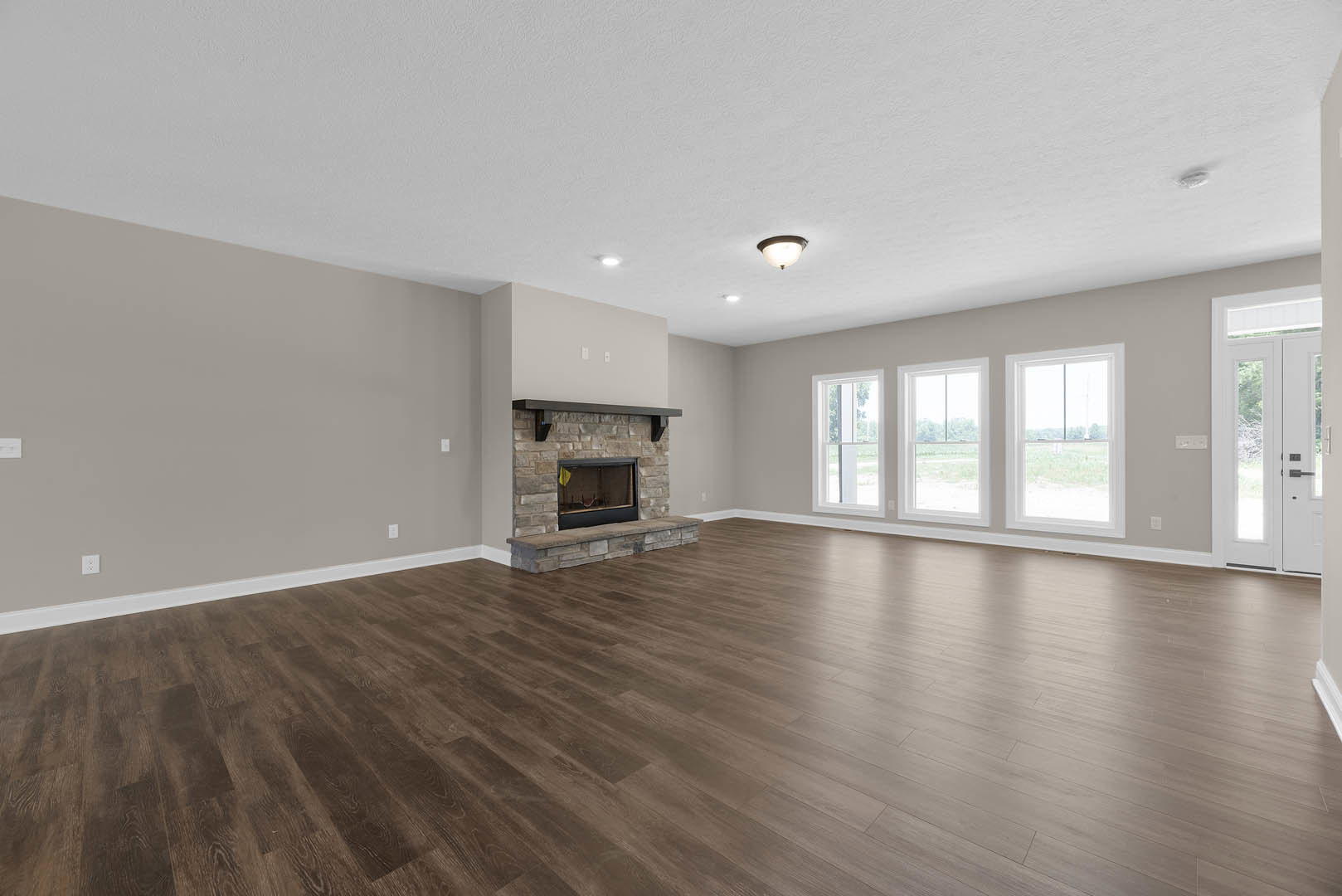 Living room with hardwood floors, white plaster walls, stone fireplace, large windows overlooking grassy yard and trees, ceiling-mounted light fixture
