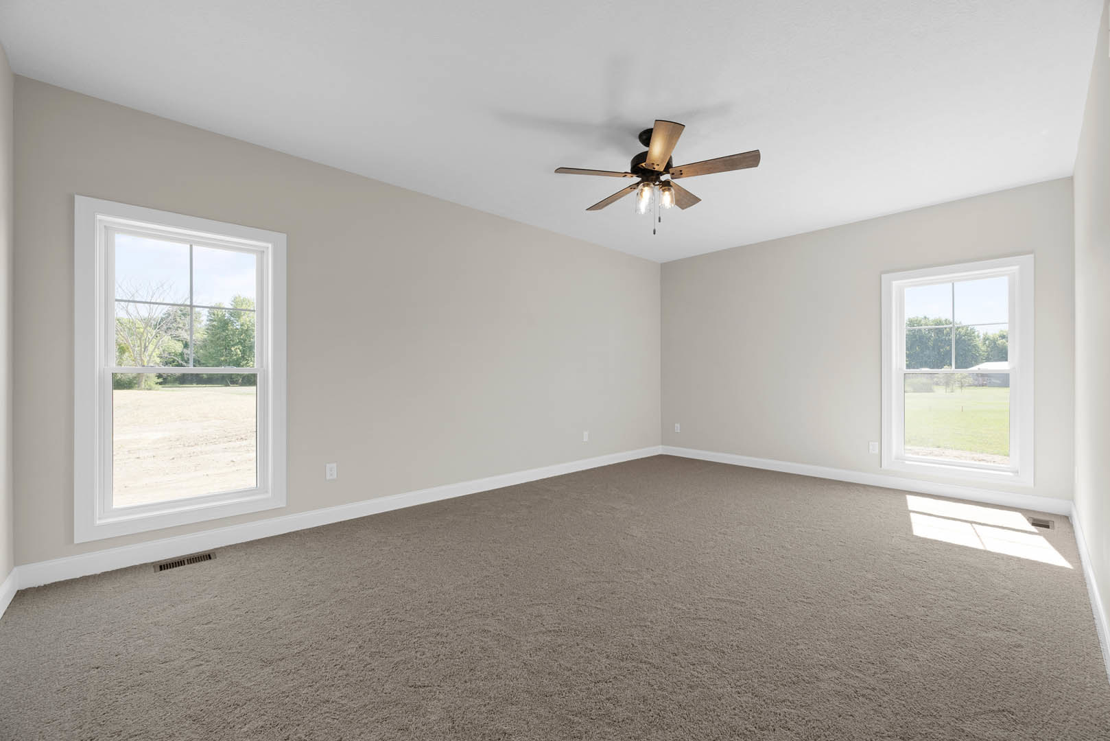 Carpeted room with white walls, ceiling fan with lights, large windows letting in natural light and offering views of a green lawn and open field