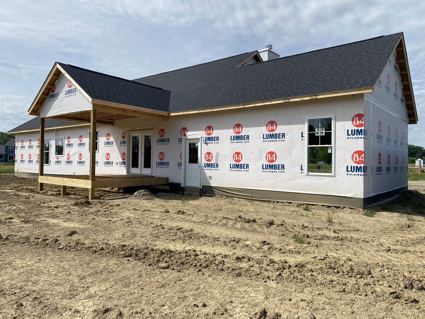 White-walled house with red and blue logos, triangular roof, window displaying a sign, white door, and patch of bare dirt in the foreground under a partly cloudy sky.