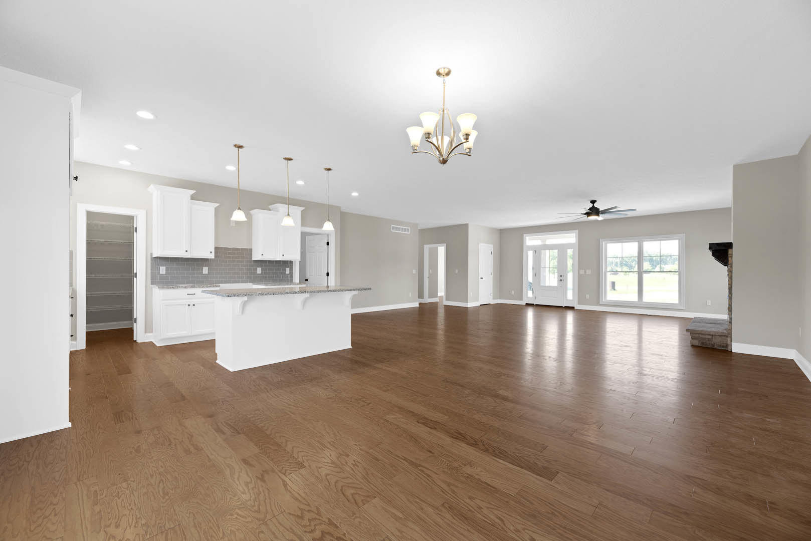 Spacious open-concept living area featuring hardwood floors, central kitchen island with marble countertop, white walls, modern chandelier, and large windows offering outdoor views