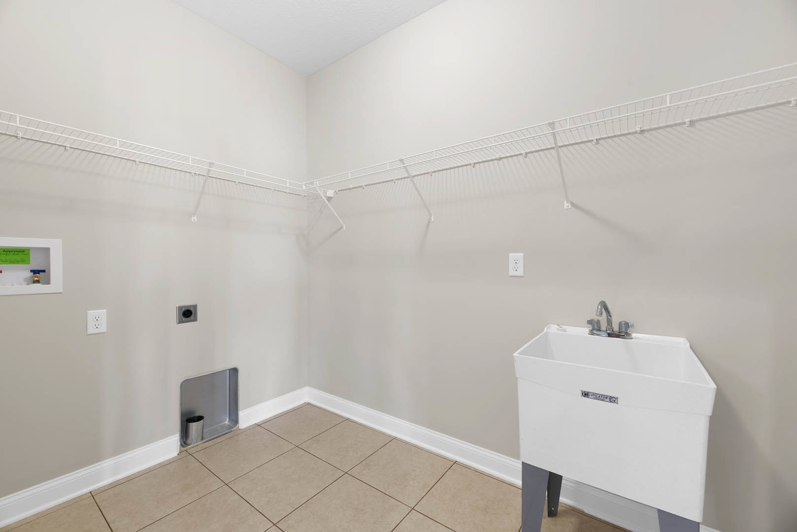 Laundry room with white tile flooring, white sink and silver faucet, silver trash can mounted on wall, white electrical outlet with black faces, white box with green label, plaster
