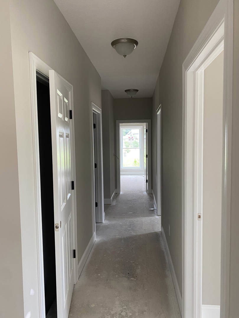 Hallway with white plaster walls, white doors featuring black handles, concrete flooring illuminated by wall light fixture, window offering view of neighboring house