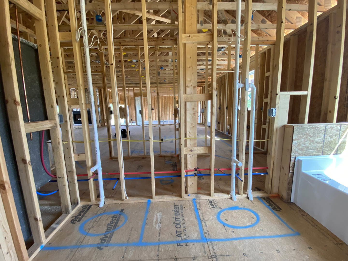 Exposed wood framing and beams in a house under construction, blue chalk lines marked on subfloor, white drywall with sunlight streaming through window, blue circle on gray