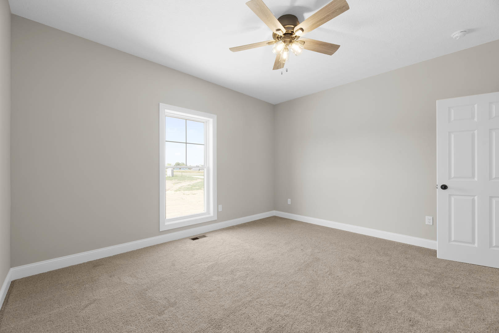 Carpeted room with white walls, ceiling fan with lights, white-framed window overlooking a grassy field, and a white paneled door