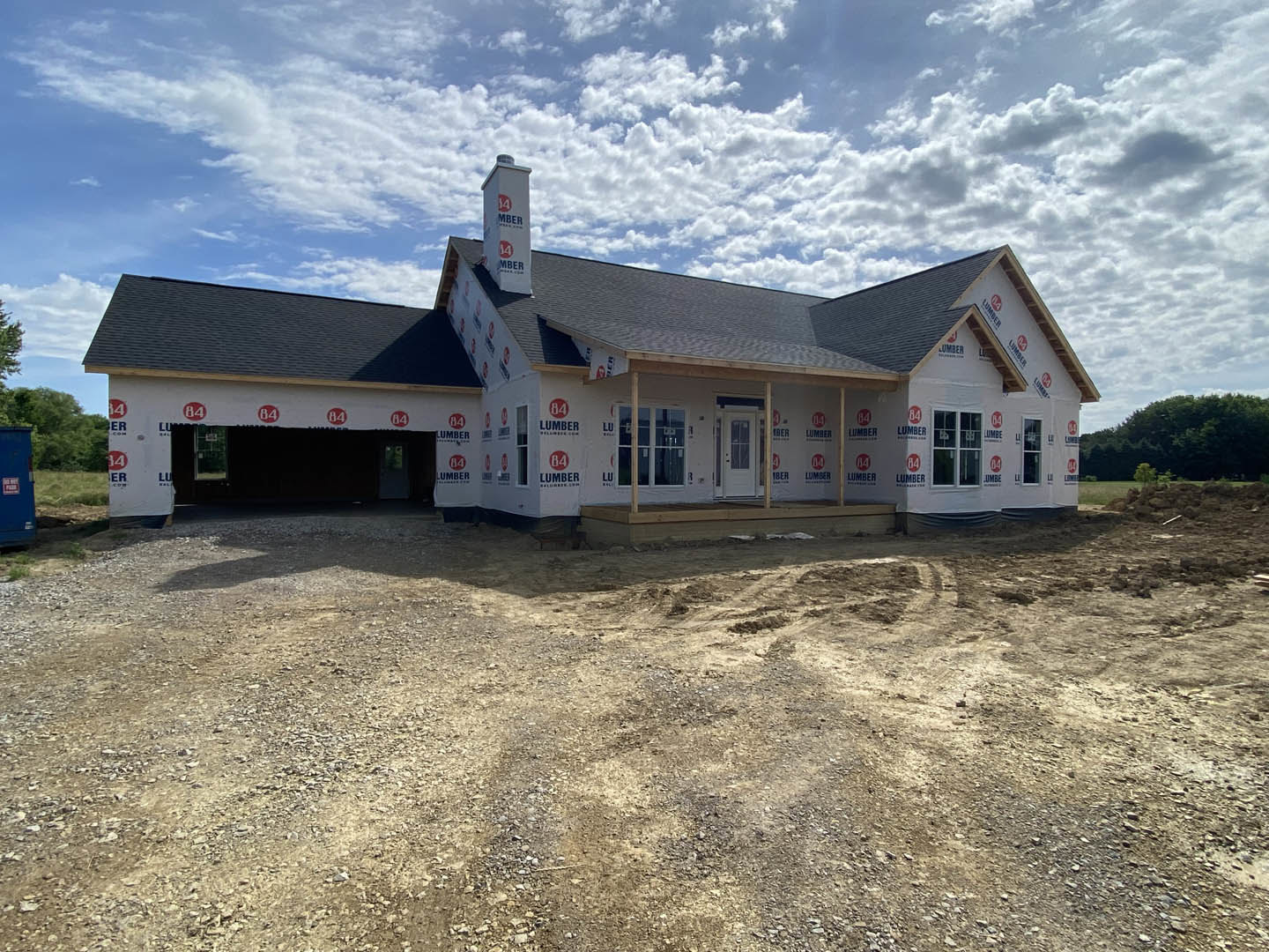 Partially built house with exposed framing and chimney, surrounded by dirt road and open ground, under a cloudy sky