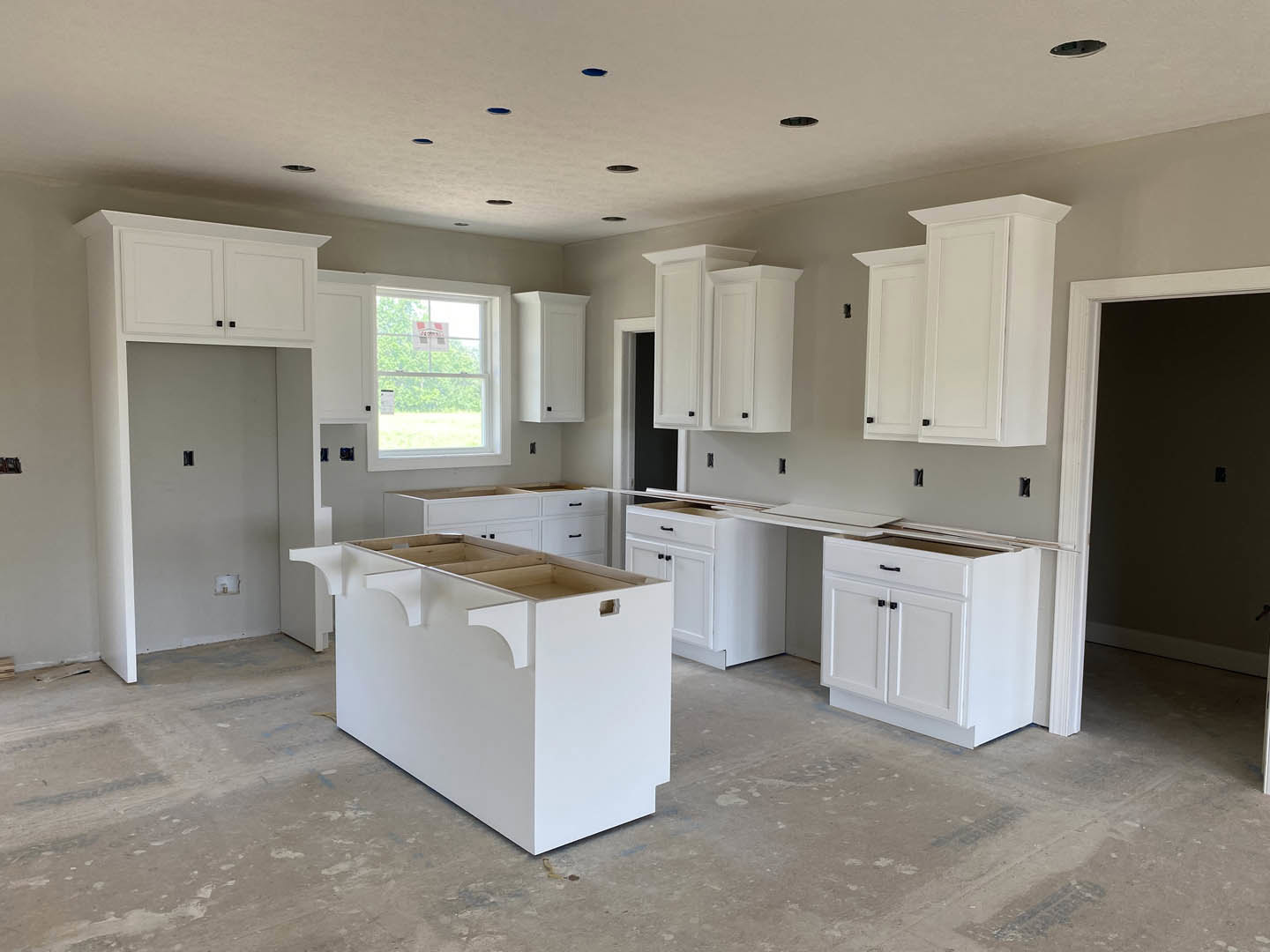 White kitchen with shaker cabinets, black handles, white countertops, central island with open shelving, stainless steel sink, window displaying a sign, light walls, and wood