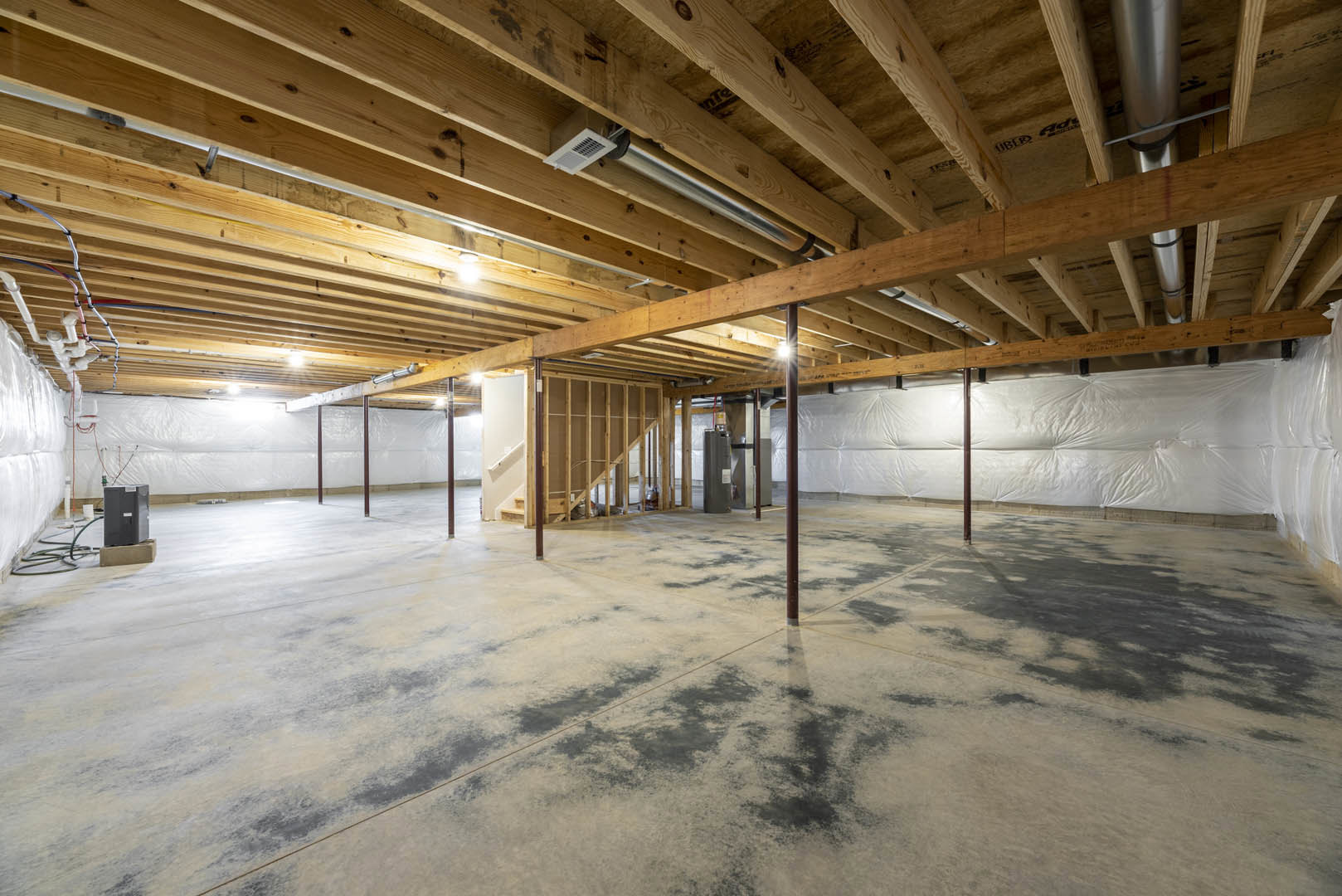 Open room with exposed wooden ceiling beams, concrete floor with black stains, staircase, recessed ceiling lights, white vent, black metal box with white label, and close-up of