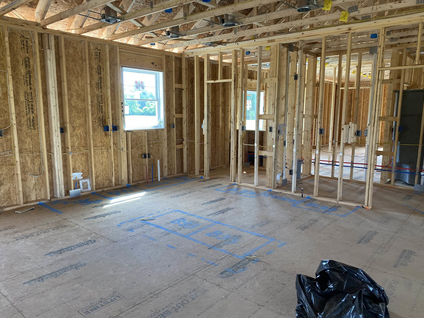 Room under construction with exposed wood framing, window displaying a sign, floor marked with blue tape, black plastic bag on ground, ceiling beams, and a blue handle on a white