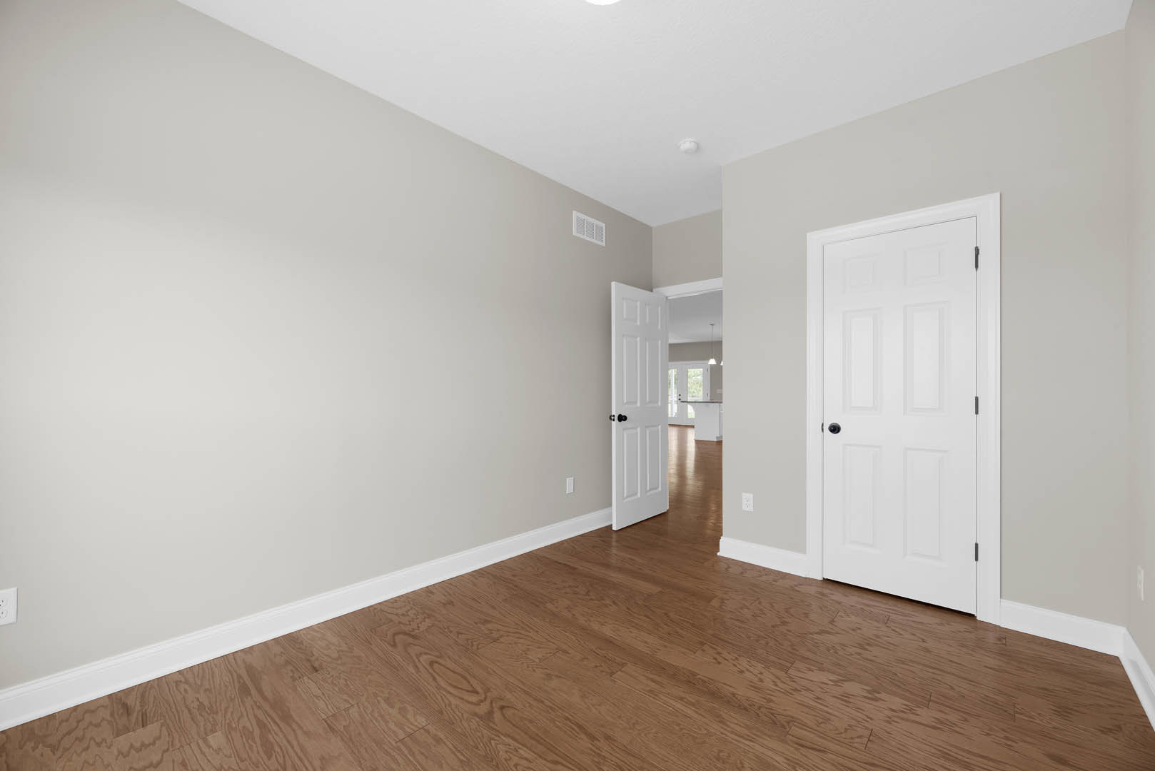 Room with white paneled doors featuring black hardware, hardwood flooring, white walls, and a white wall vent