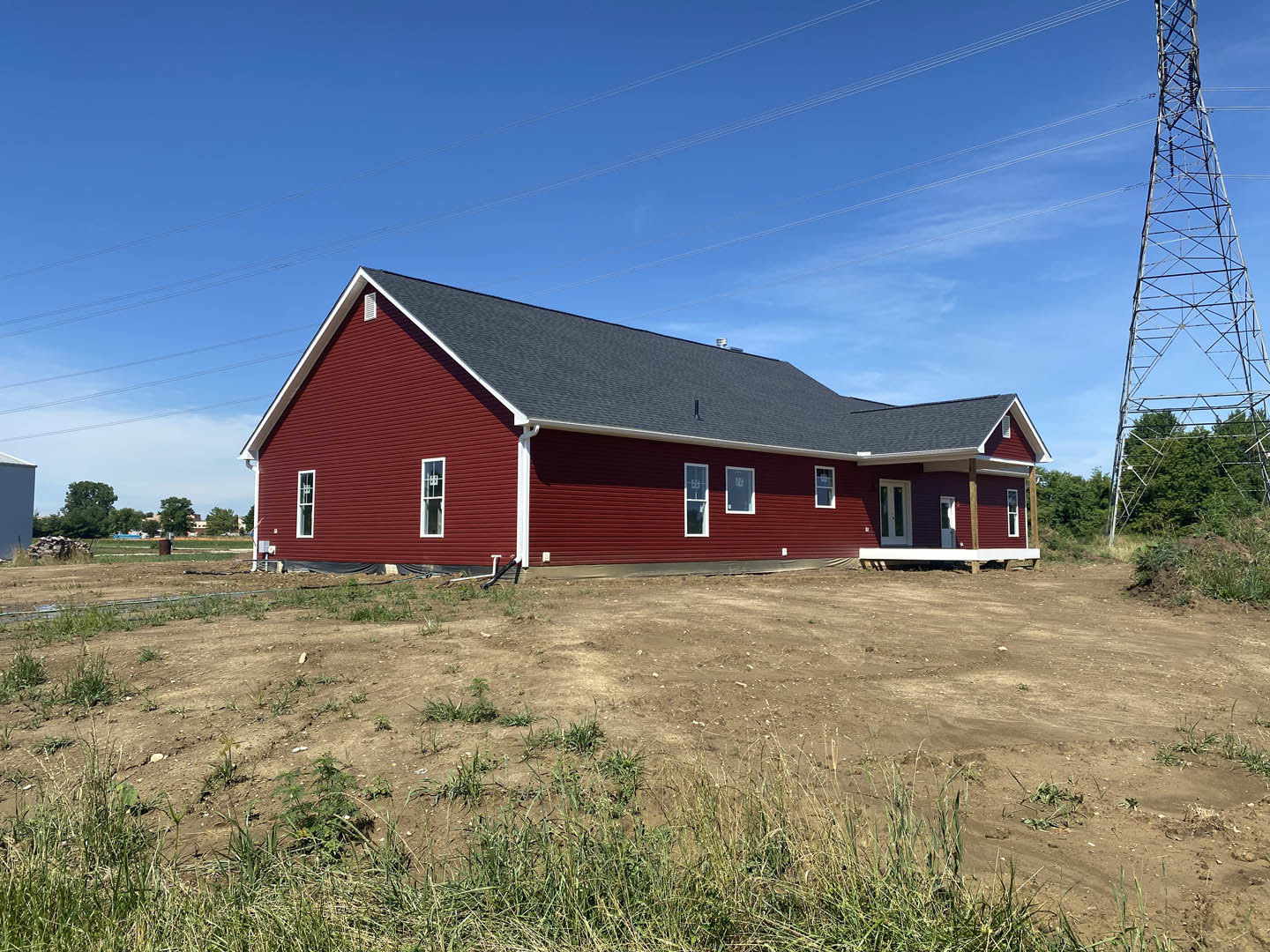 Red house with white trim and gabled roof, surrounded by dirt field with patches of grass, power lines overhead, metal tower nearby, rural landscape with trees and cloudy sky.