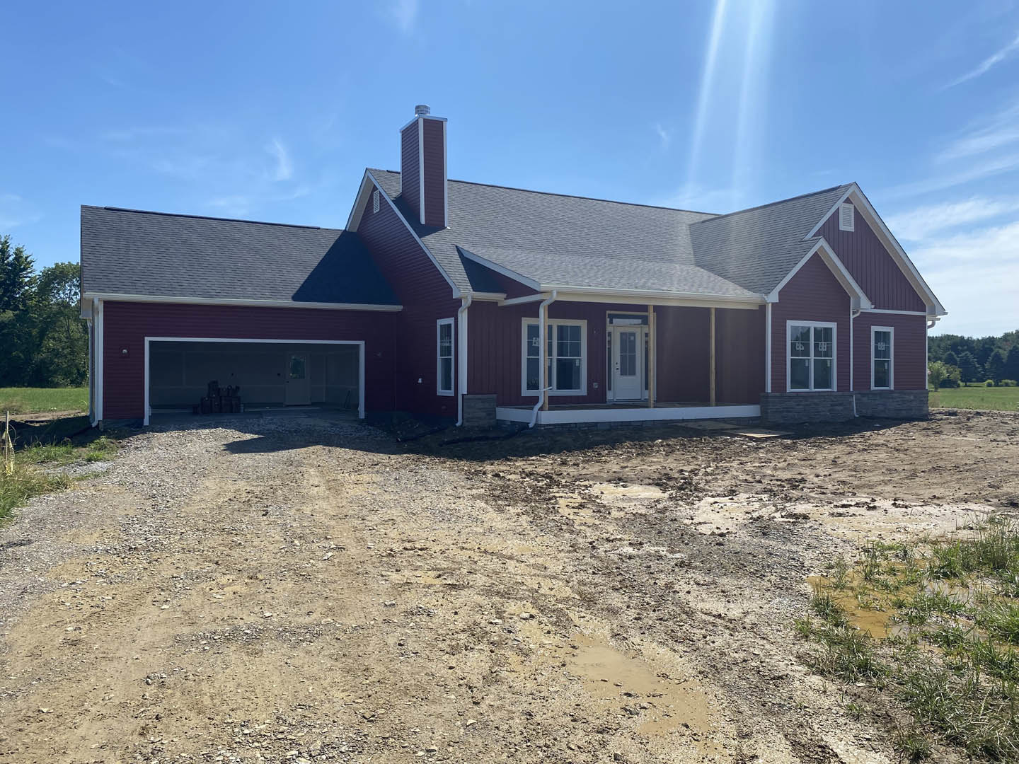 Two-story house under construction with white garage door, glass-paneled front door, exposed dirt and gravel driveway, large windows, and unfinished landscaping.