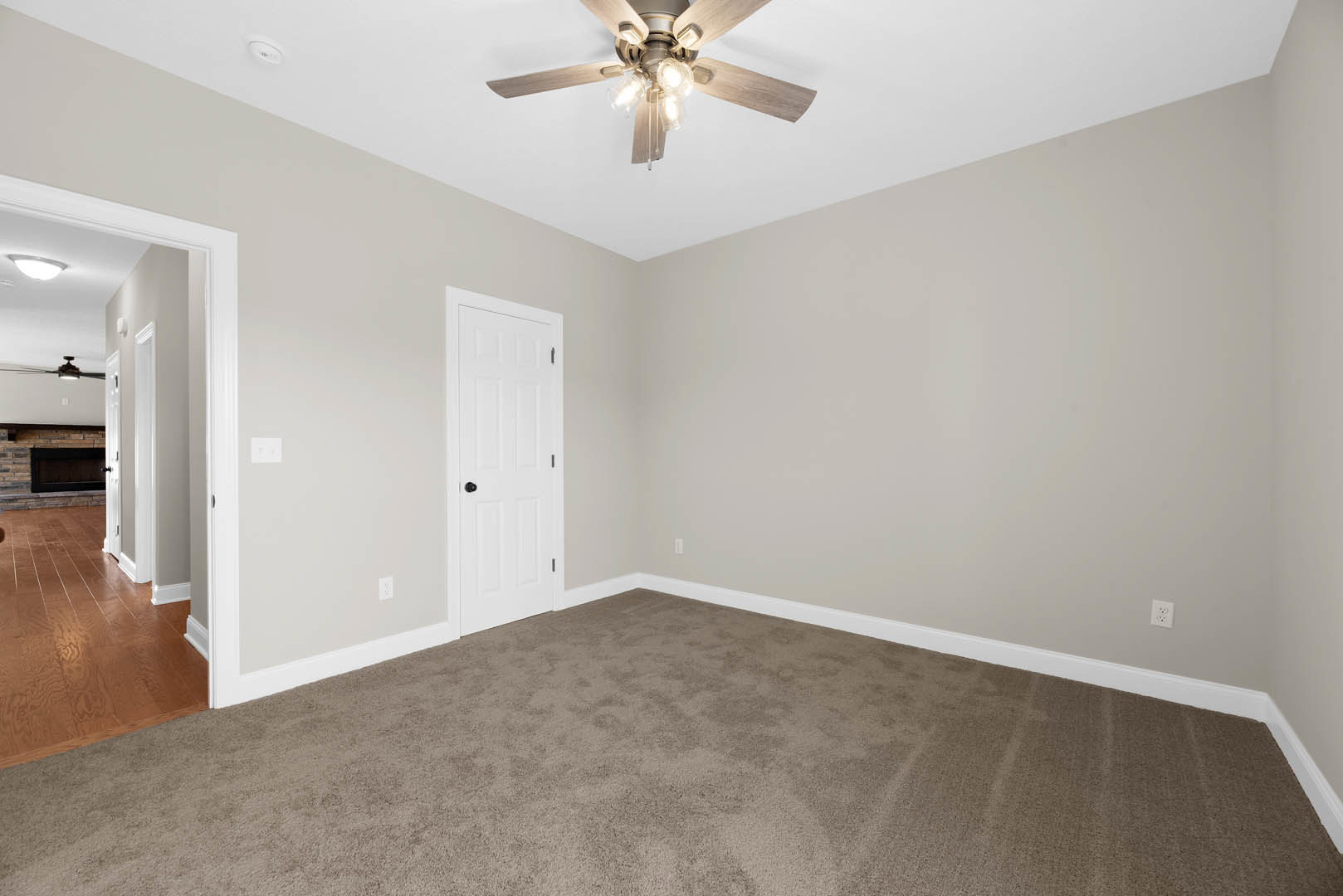 Ceiling fan with lights above carpeted room, white walls, white door with black knob, hardwood floor visible in adjacent hallway