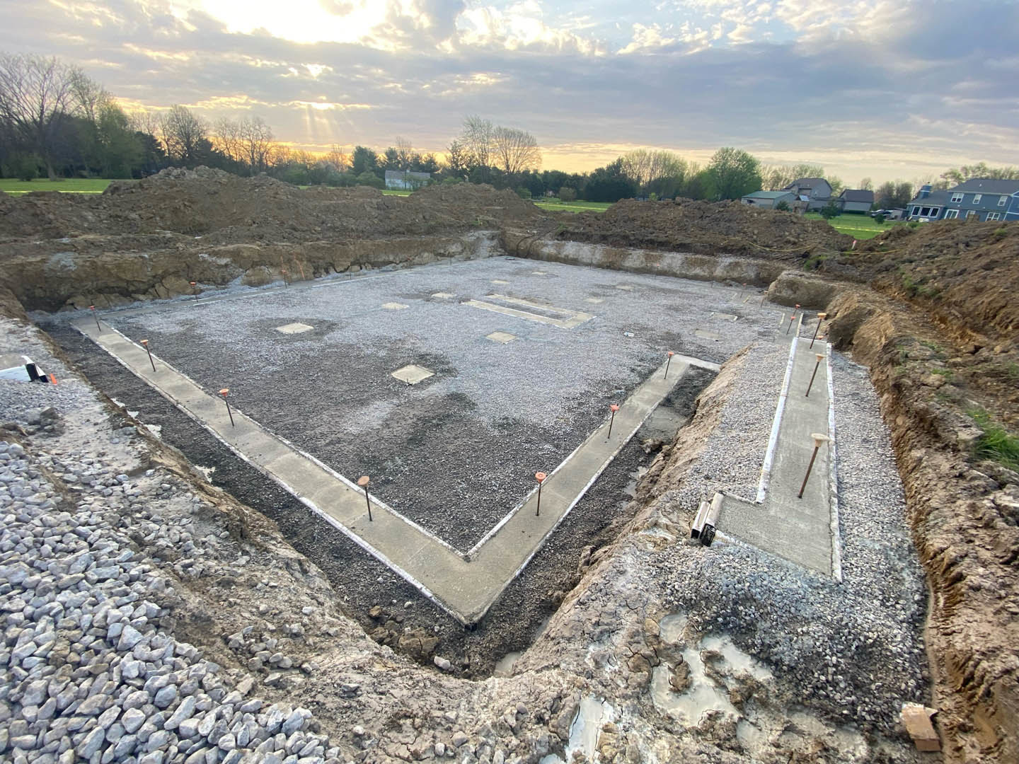 Square concrete foundation at residential construction site, surrounded by dirt and scattered rocks, with cloudy sky and trees in background