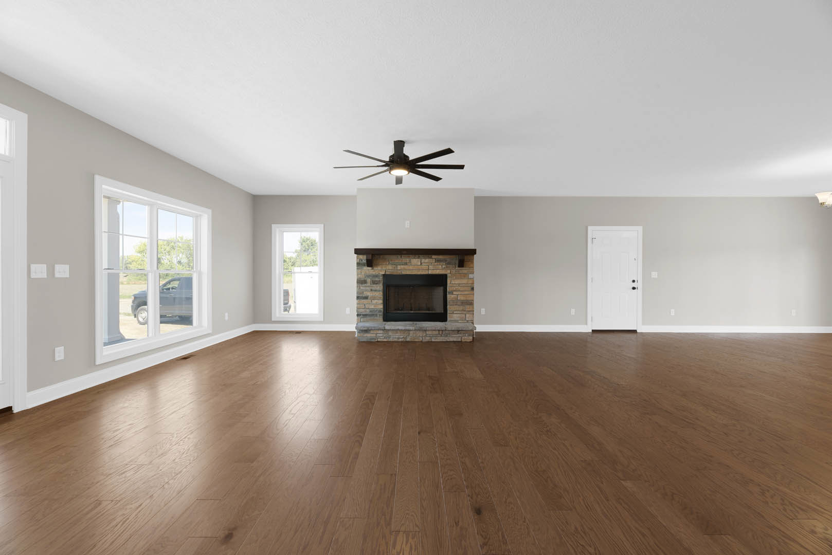 Spacious living room featuring hardwood floors, black-framed glass fireplace, ceiling fan with light, and white door with black handles
