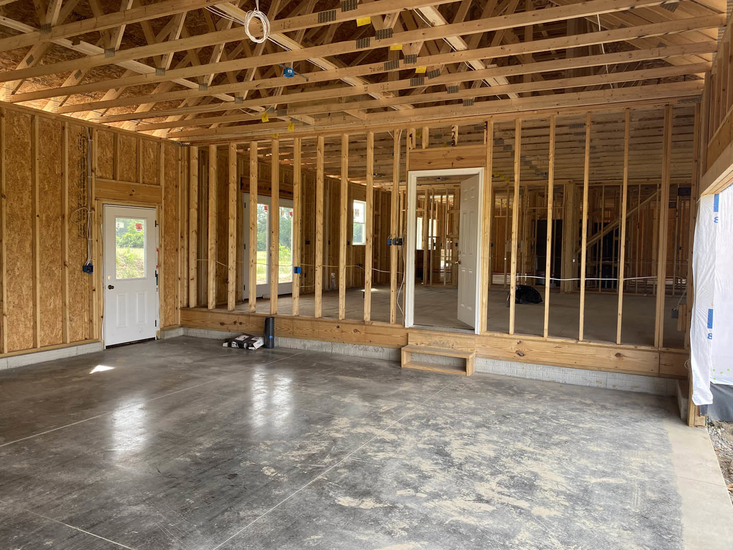 Wood-framed room with exposed roof trusses featuring yellow tags, white door with window, gray floor and wooden steps, visible wiring, and unfinished walls.