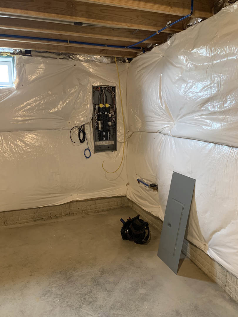 Room under renovation with plastic sheeting covering walls and floor, electrical box with exposed wires, white door with protective cover, black backpack on floor, and construction