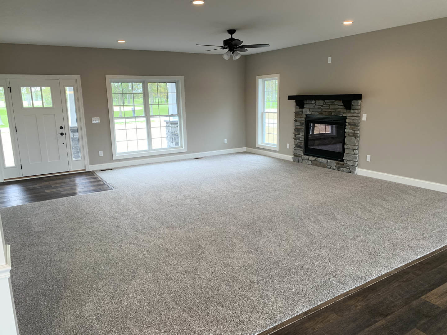 Living room with brick fireplace, ceiling fan with light fixture, laminate wood flooring, plaster walls, window overlooking lawn, and white door with glass panels