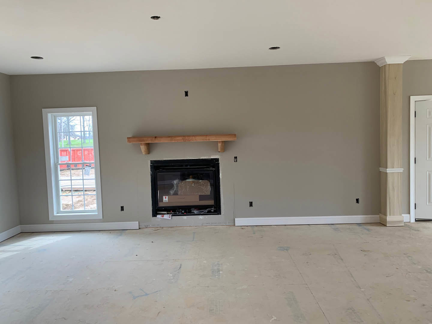 Living room with concrete floor, white plaster walls, wooden ceiling beam, fireplace, large window with white frame, and white door with black hinges
