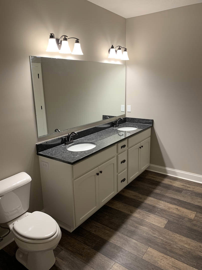 Bathroom with expansive wall mirror above double sinks, marble countertop, chrome faucets, and modern light fixtures; partial view of toilet and tiled floor.