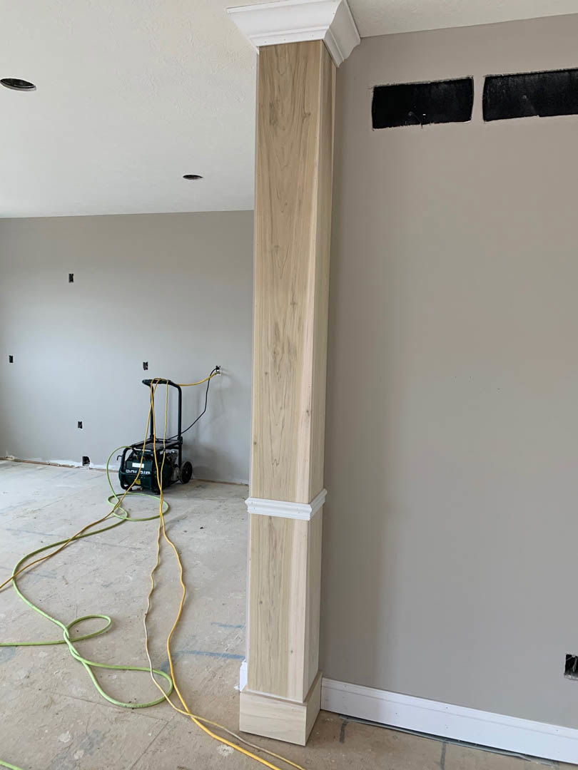 Wood pillar with natural grain standing beside a white plaster wall, black square accent on surface, wooden frame detail, green and yellow hose on light flooring, ceiling and