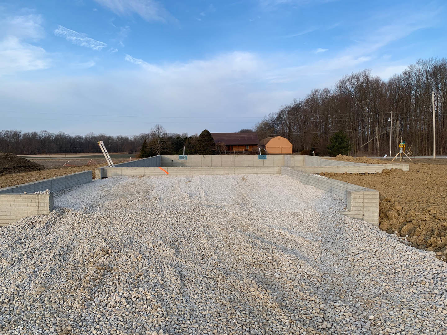 Wood-framed house under construction with exposed beams, pile of gravel in foreground, ladder leaning against unfinished wall, mature trees and blue sky with scattered clouds in