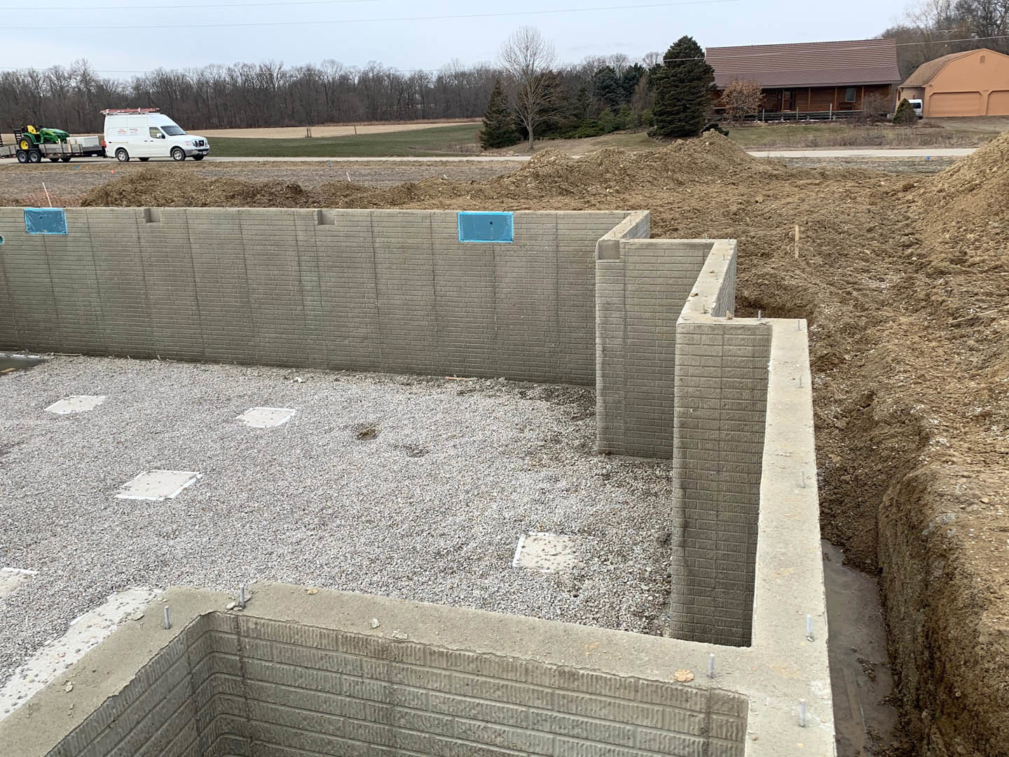 Partially constructed house with exposed foundation and dirt pit in foreground, white van parked on road, tractor on trailer, completed home with shingle roof in background, trees