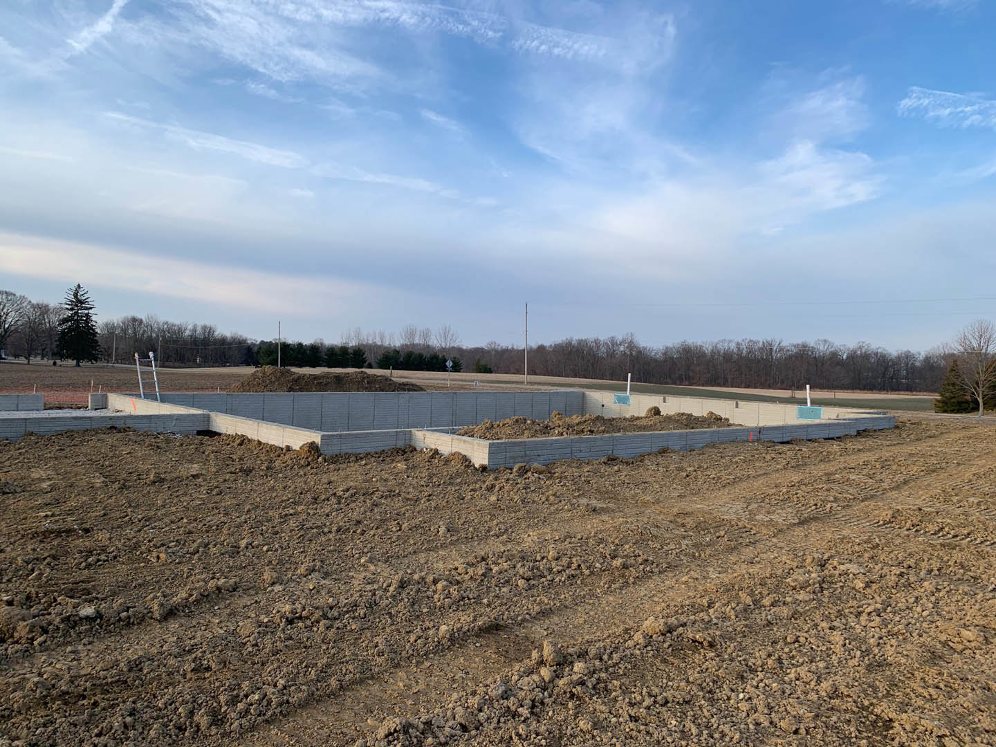 Dirt construction site bordered by a large concrete wall, scattered trees in the background, blue sky with clouds overhead