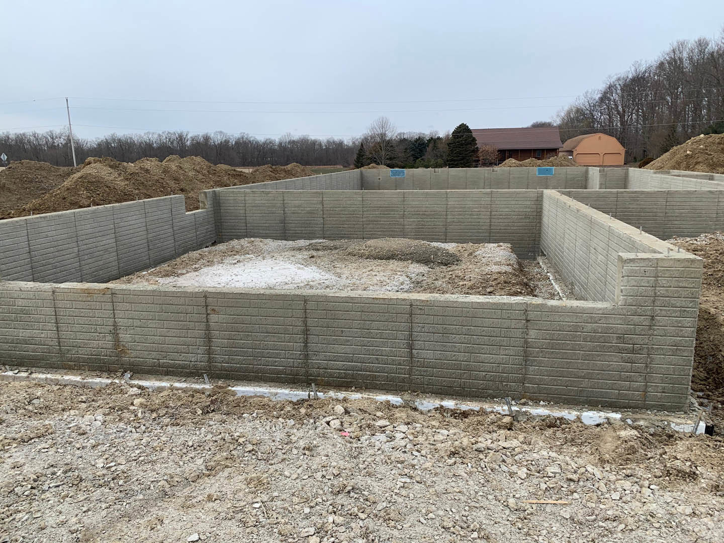 Concrete foundation surrounded by piles of dirt and rocks, partially constructed wall, brown tent in open field, blue sky with scattered clouds in background.