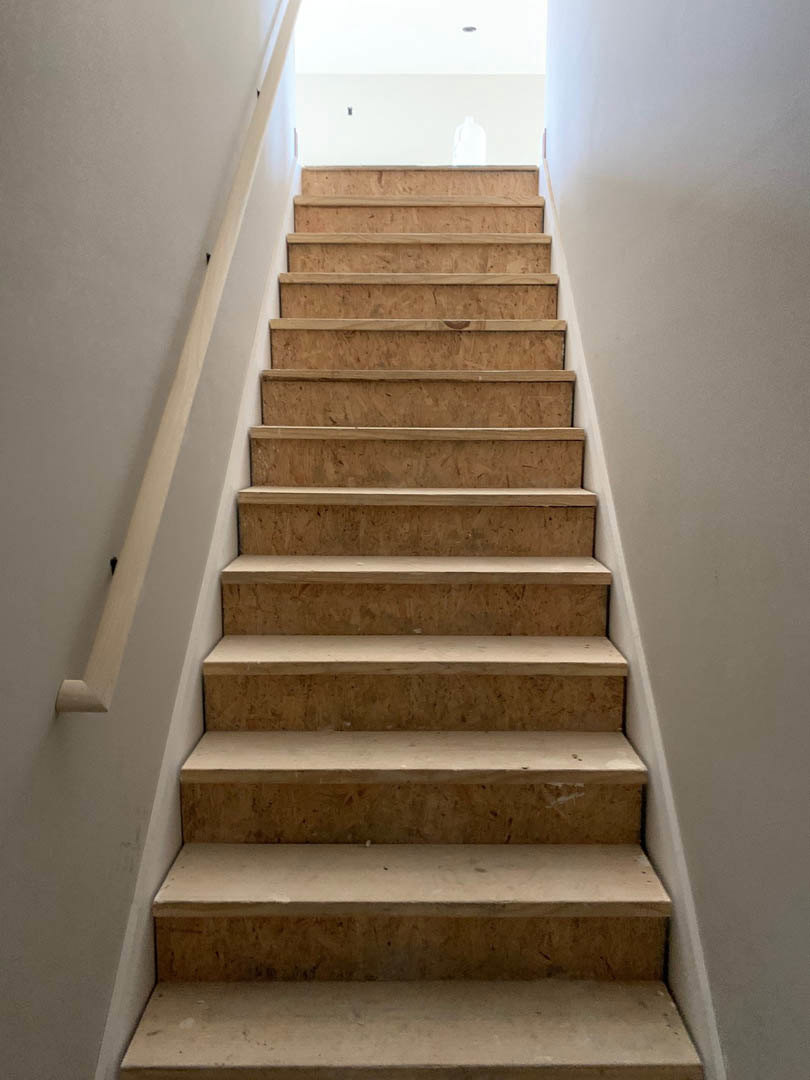 Wooden staircase with white handrail, sunlight streaming through nearby window onto light-colored walls, close-up of built-in shelf and smooth white surface.