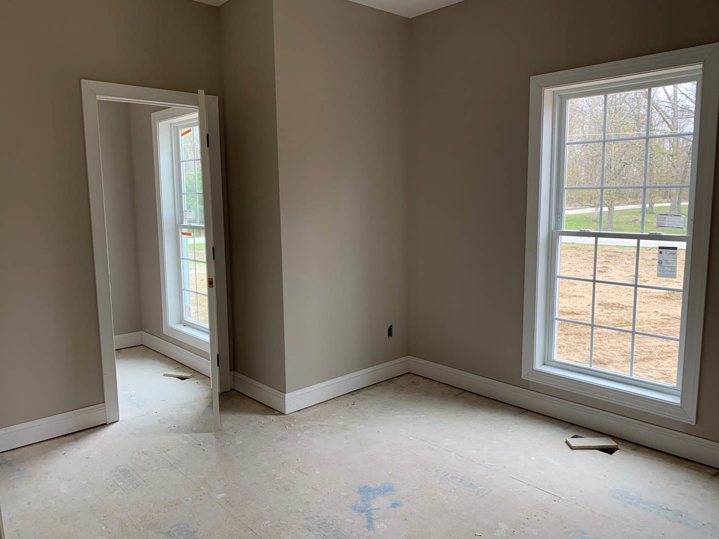 White-walled room with two rectangular windows, a door, and a dirty floor featuring patches of blue paint; sunlight streams in, illuminating the unfinished interior.