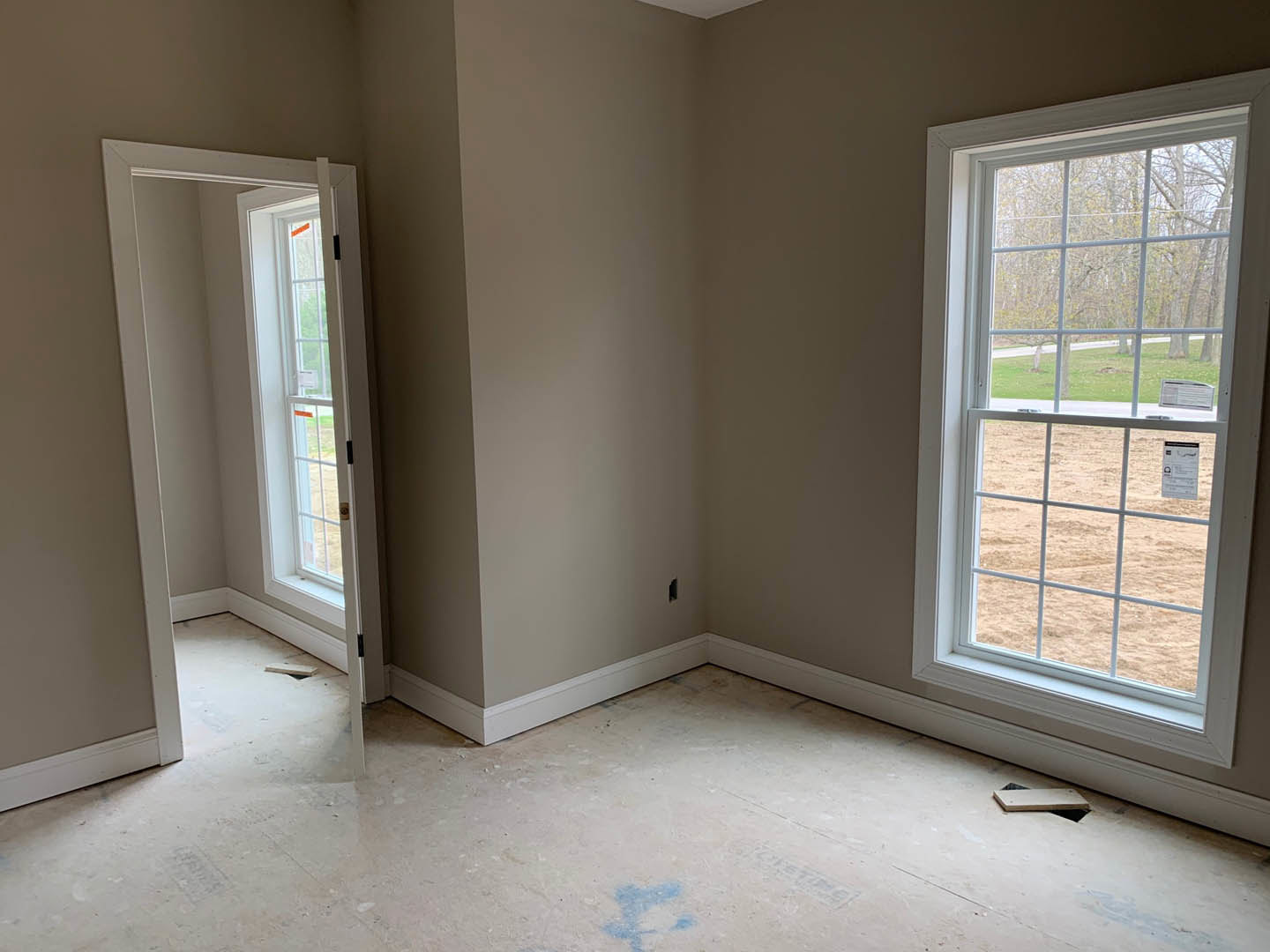 Room with two large windows and a door, white plaster walls, window blinds, and a floor with blue paint stains; daylight streams in, revealing a field outside.