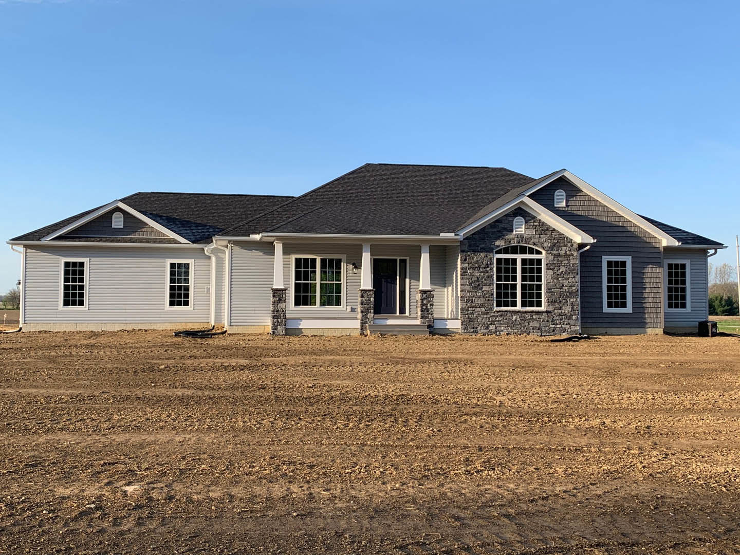 Two-story farmhouse with black roof, white siding, and multi-pane windows, set beside a large dirt field under a clear sky