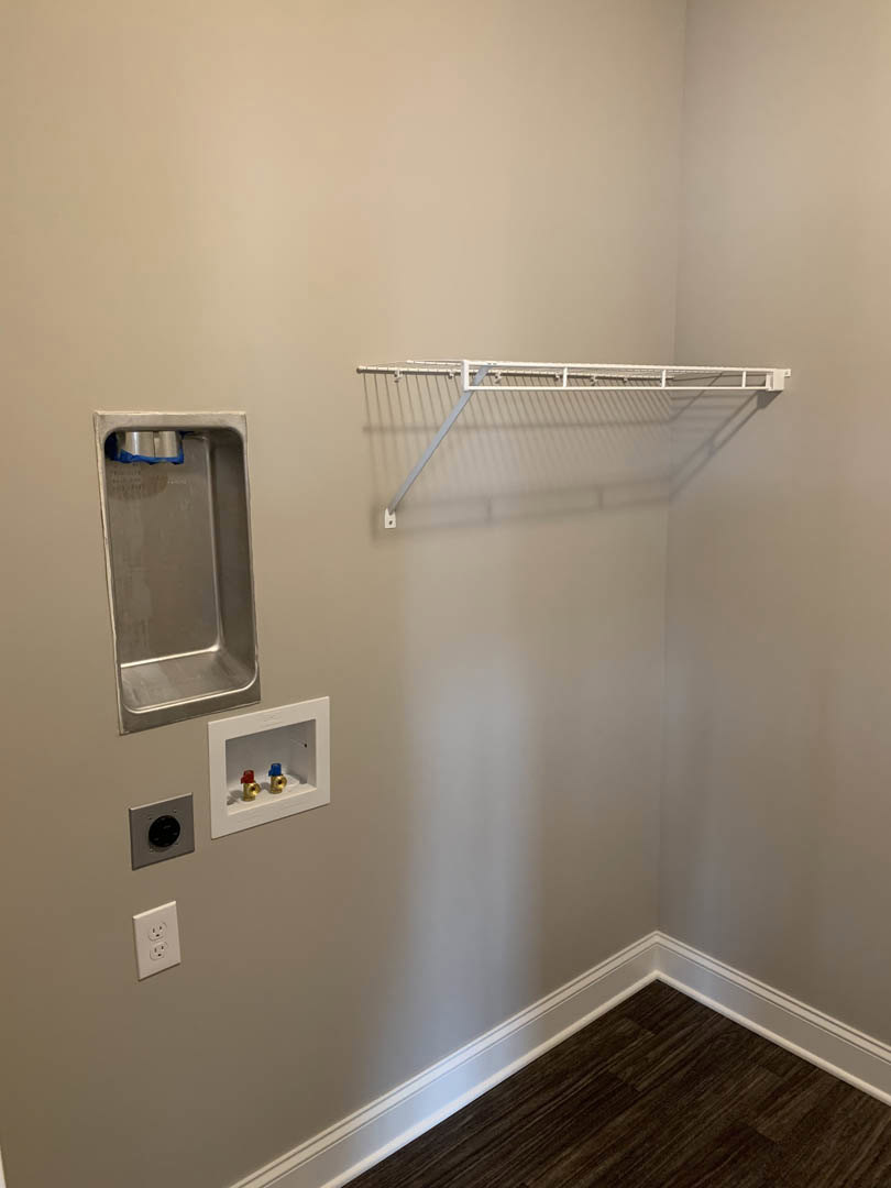 White metal rack mounted on plaster bathroom wall above dark wood floor and white baseboard, brass pipes and electrical outlet visible nearby.