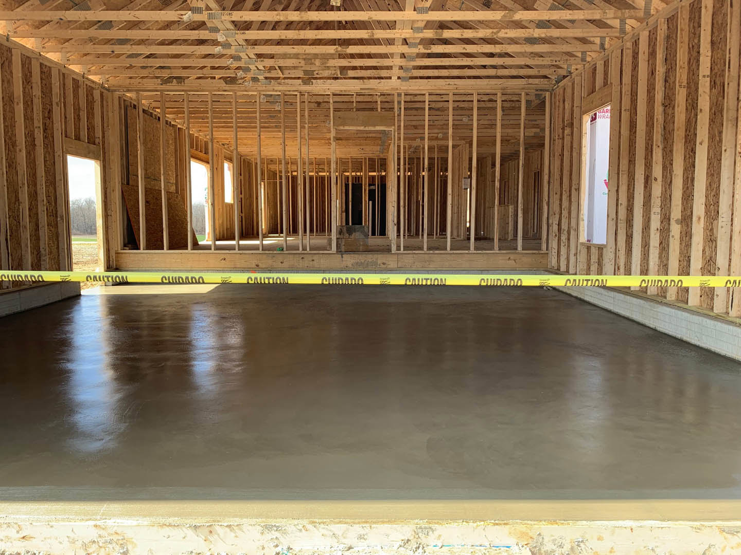 Concrete floor under construction with yellow caution tape, exposed wooden ceiling beams, white wall featuring pink and green sticker, large window revealing tree outside