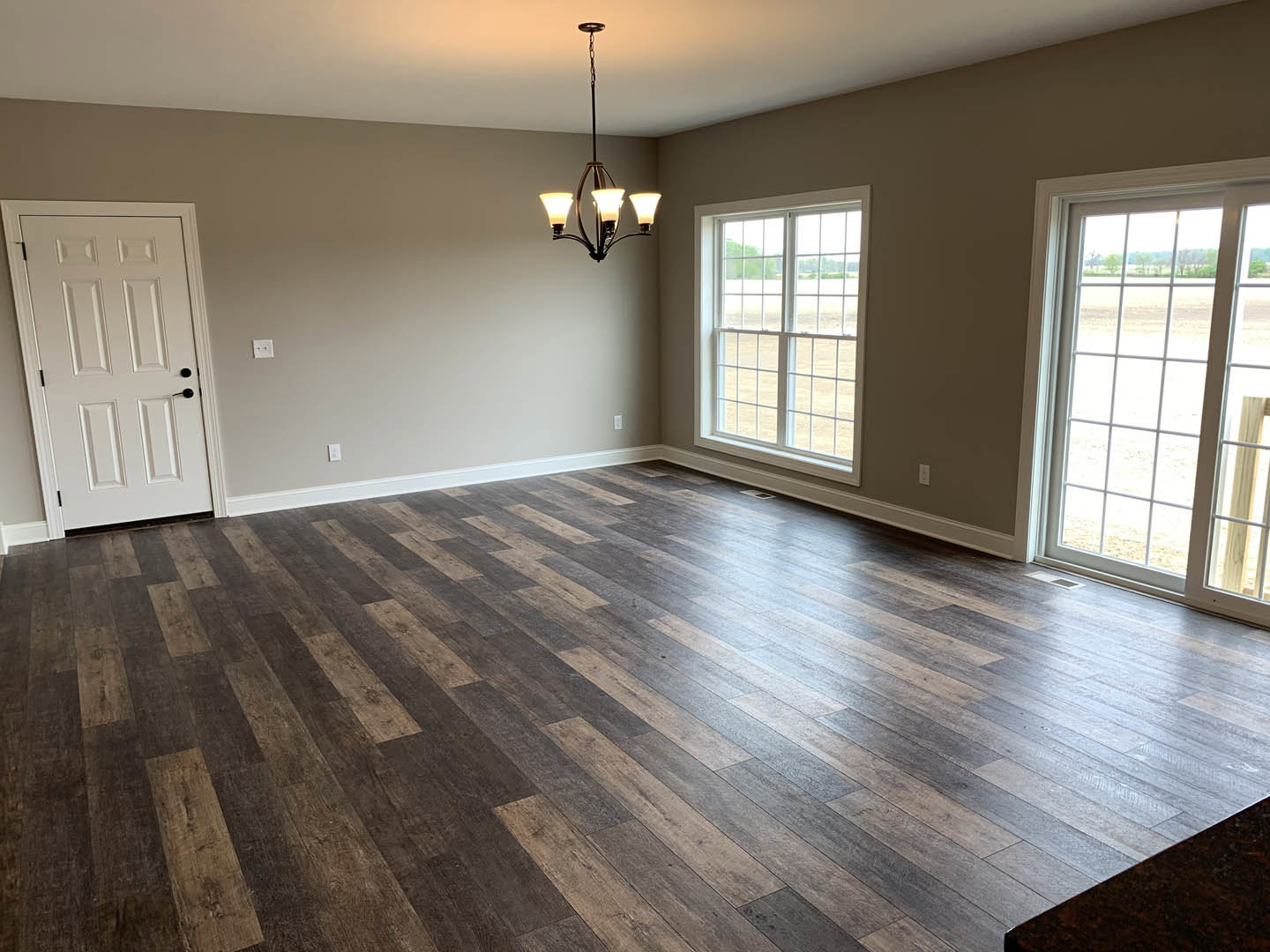 Wood flooring in a spacious room, white door with black handles, large window overlooking a grassy field, ornate chandelier hanging from the ceiling.