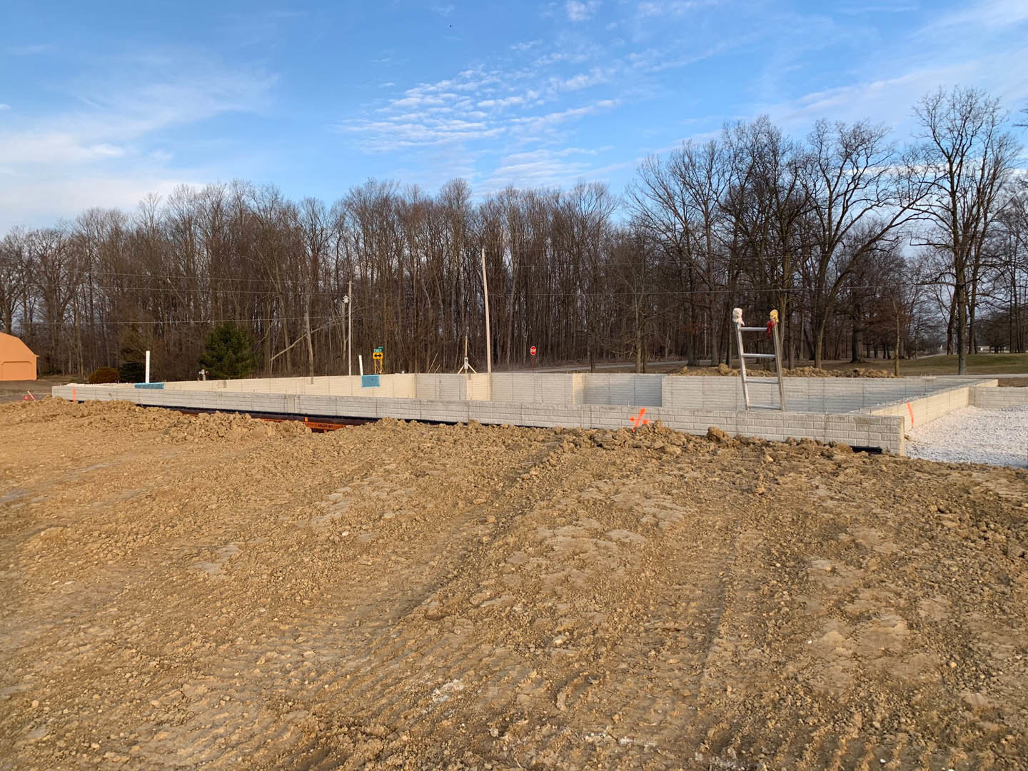 Dirt construction site bordered by a metal fence, orange cones scattered across the ground, partially built wall in foreground, trees and blue sky with clouds in background